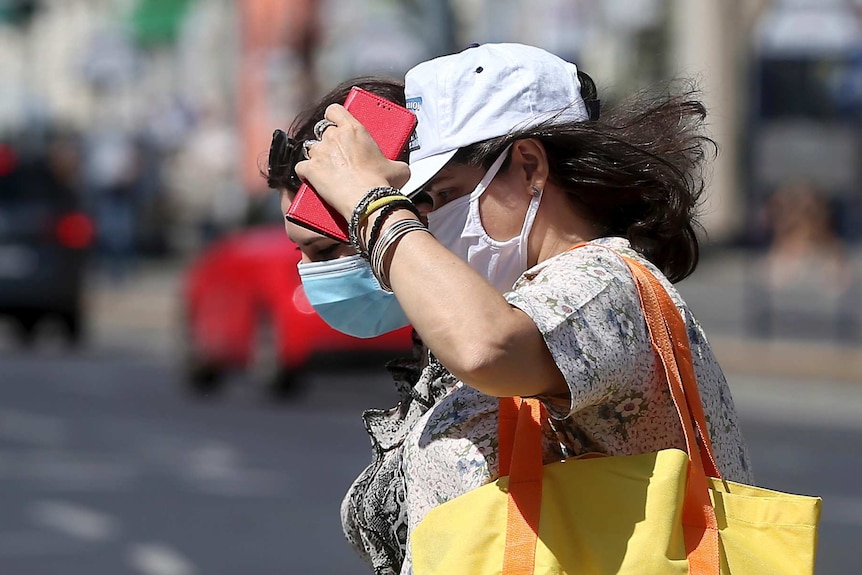 Two women wearing face masks walk across a street in bright sunlight.