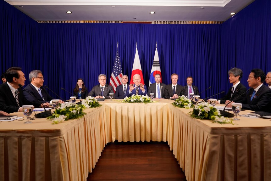 Joe Biden sits at a table and meets with Fumio Kishida and Yoon Suk Yeol.