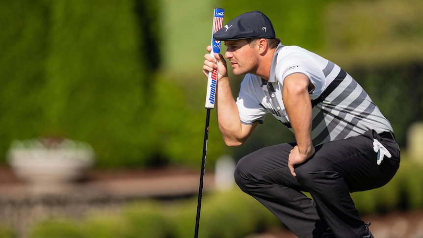 Bryson DeChambeau crouches with his putter on the green.