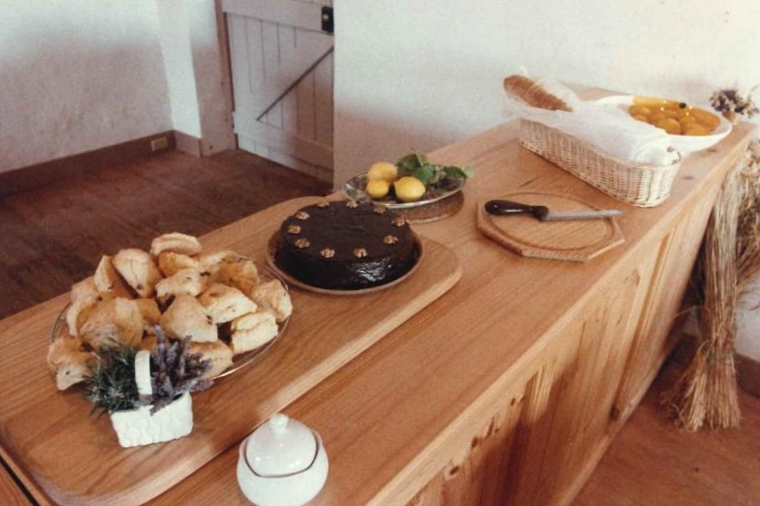A wooden bench lined with scones, a chocolate covered cake, a bread loaf, lemons, lavender and a chopping board.