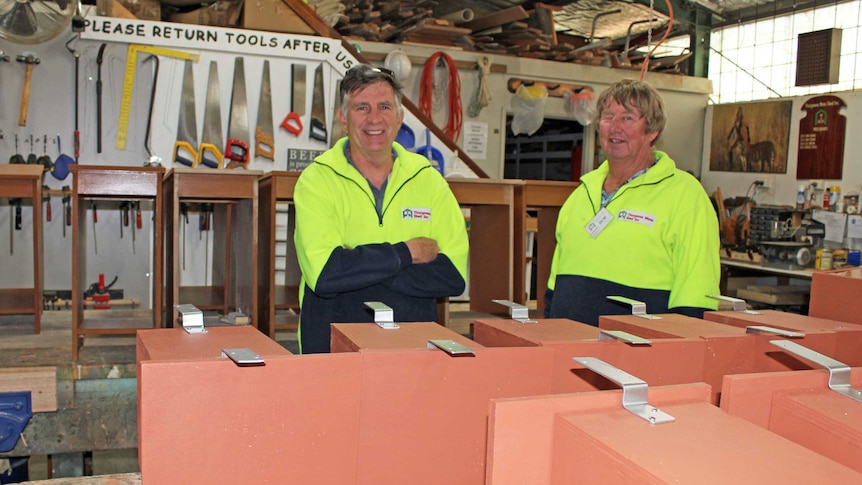 Two men are wearing yellow high vis jumpers are standing in a wood work shop behind a table of nest boxes