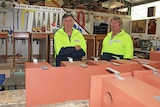 Two men are wearing yellow high vis jumpers are standing in a wood work shop behind a table of nest boxes