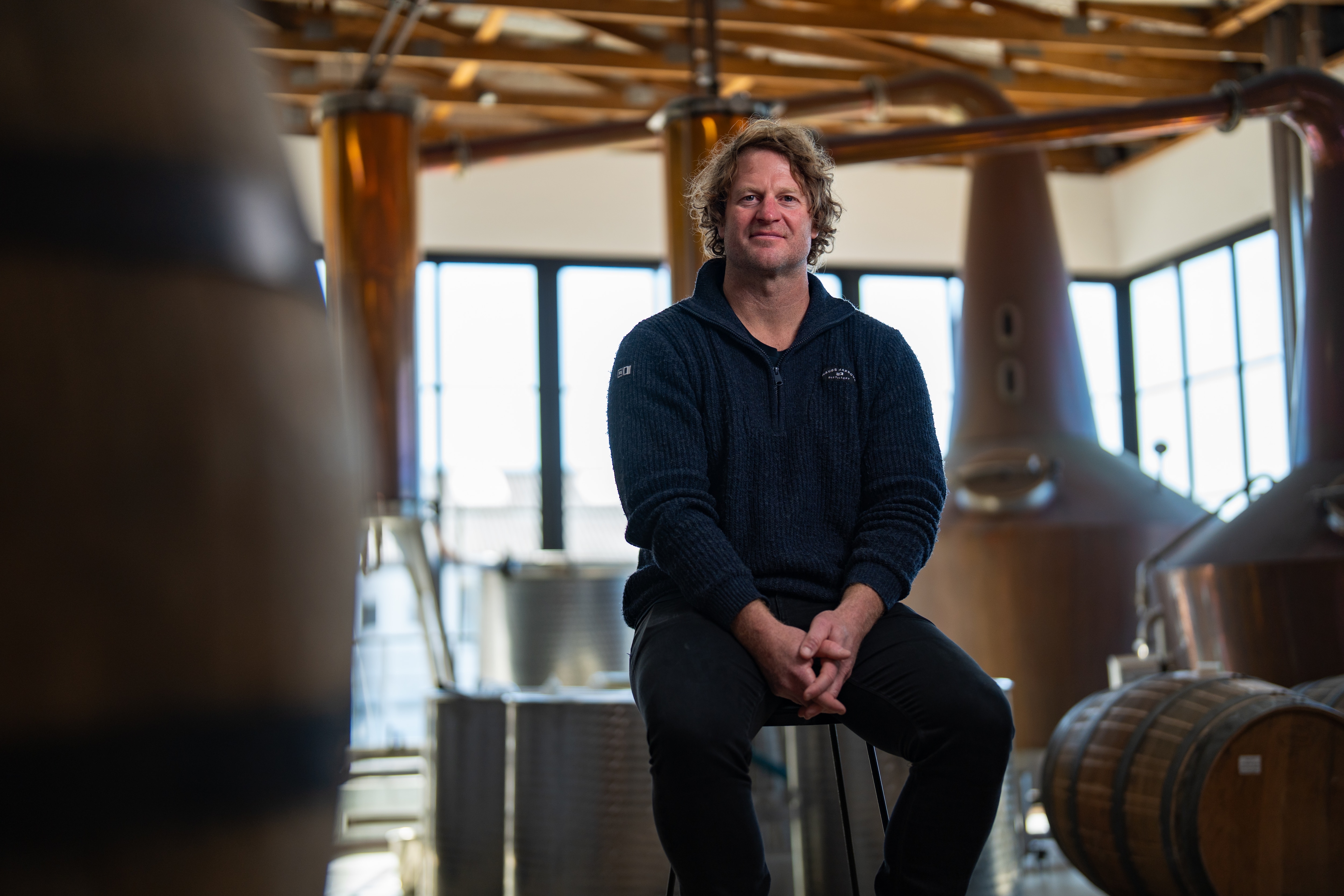 A man with curly hair sits surrounded by large copper distilling equipment
