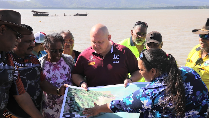 Aboriginal Elders and community stand in from of a jetty under construction and look over a map spread out in front of them.