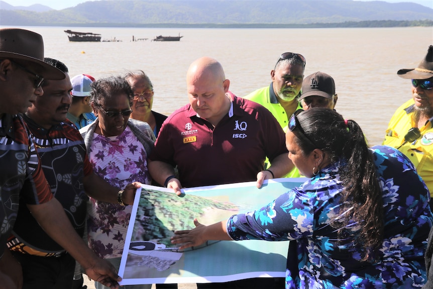 Aboriginal Elders and community stand in from of a jetty under construction and look over a map spread out in front of them.