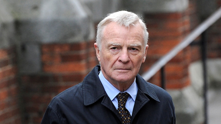 Max Mosley stands in the street wearing a dark trenchcoat.