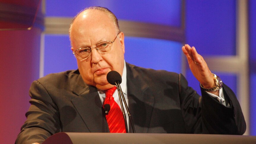 Roger Ailes gestures as he stands at a lectern with Fox written on it.