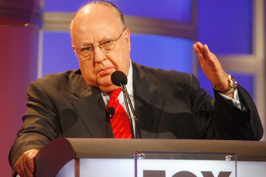 Roger Ailes gestures as he stands at a lectern with Fox written on it.
