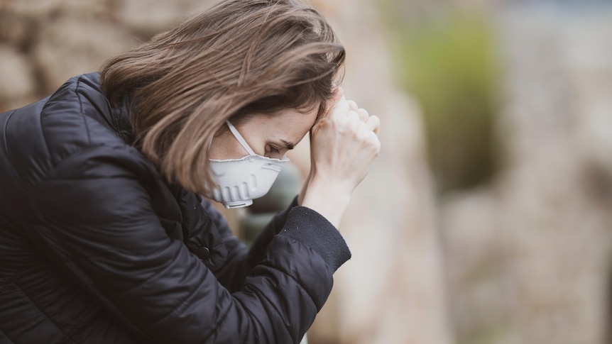 A woman in a face masks holds her head in her hands.