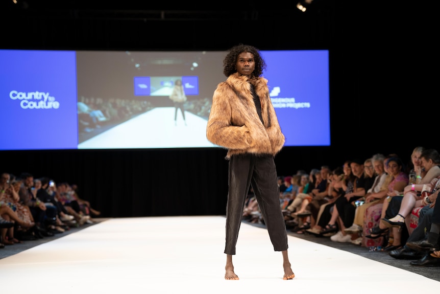 A man with curly hair and wearing a fur coat stands on a fashion runway. 