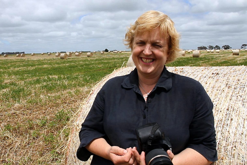 A fair-haired woman wearing a navy blue shirt and carrying her camera, stands in front of a hay bale 