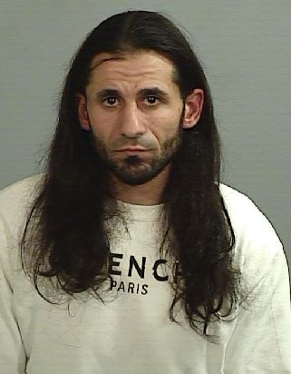 A grainy mugshot of a long-haired man with a short beard and dark features