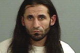 A grainy mugshot of a long-haired man with a short beard and dark features