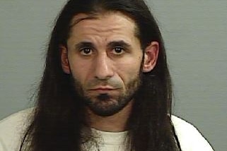 A grainy mugshot of a long-haired man with a short beard and dark features