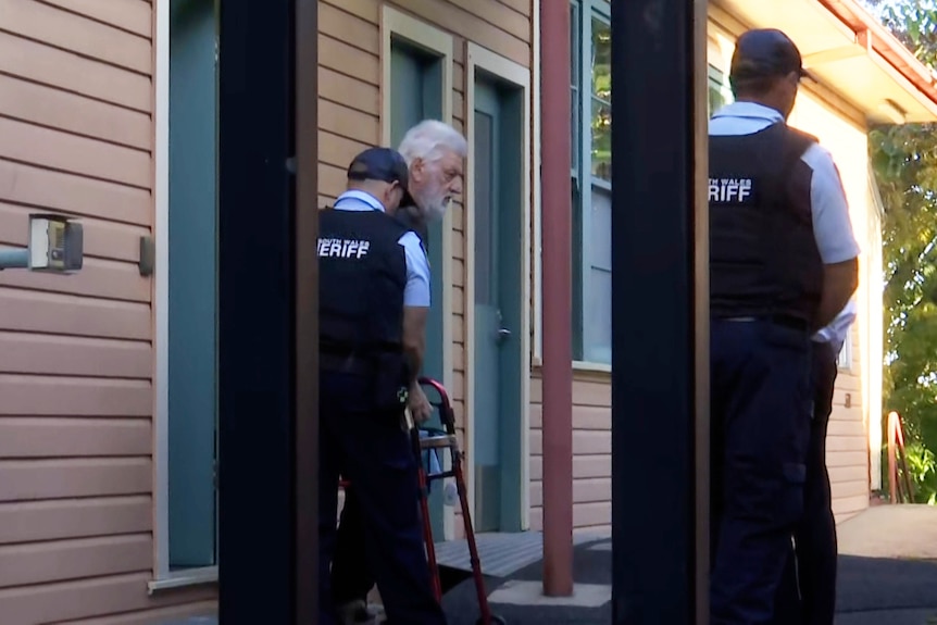Camera through a gate showing an elderly man with white hair being escorted out of a building by police