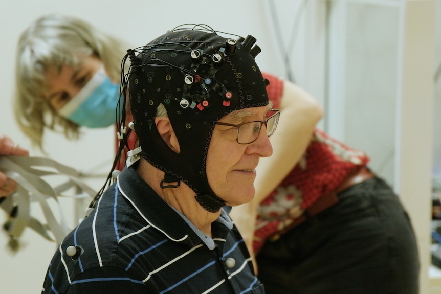 An elderly man sits in profile wearing a black helmet fitted with sensors. A researcher in a face mask works in the background. 