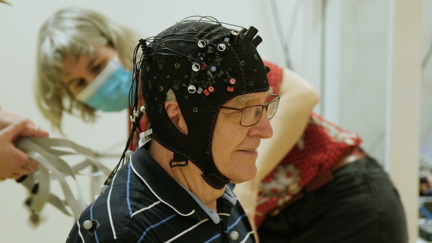 An elderly man sits in profile wearing a black helmet fitted with sensors. A researcher in a face mask works in the background. 