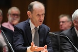 Shayne Elliott seated in front of a microphone, wearing a dark suit and gesturing with his hands.