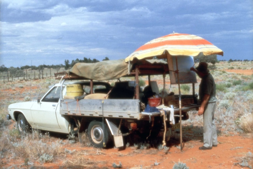 A man paints under an orange-and-yellow umbrella rigged up over the back of a ute.