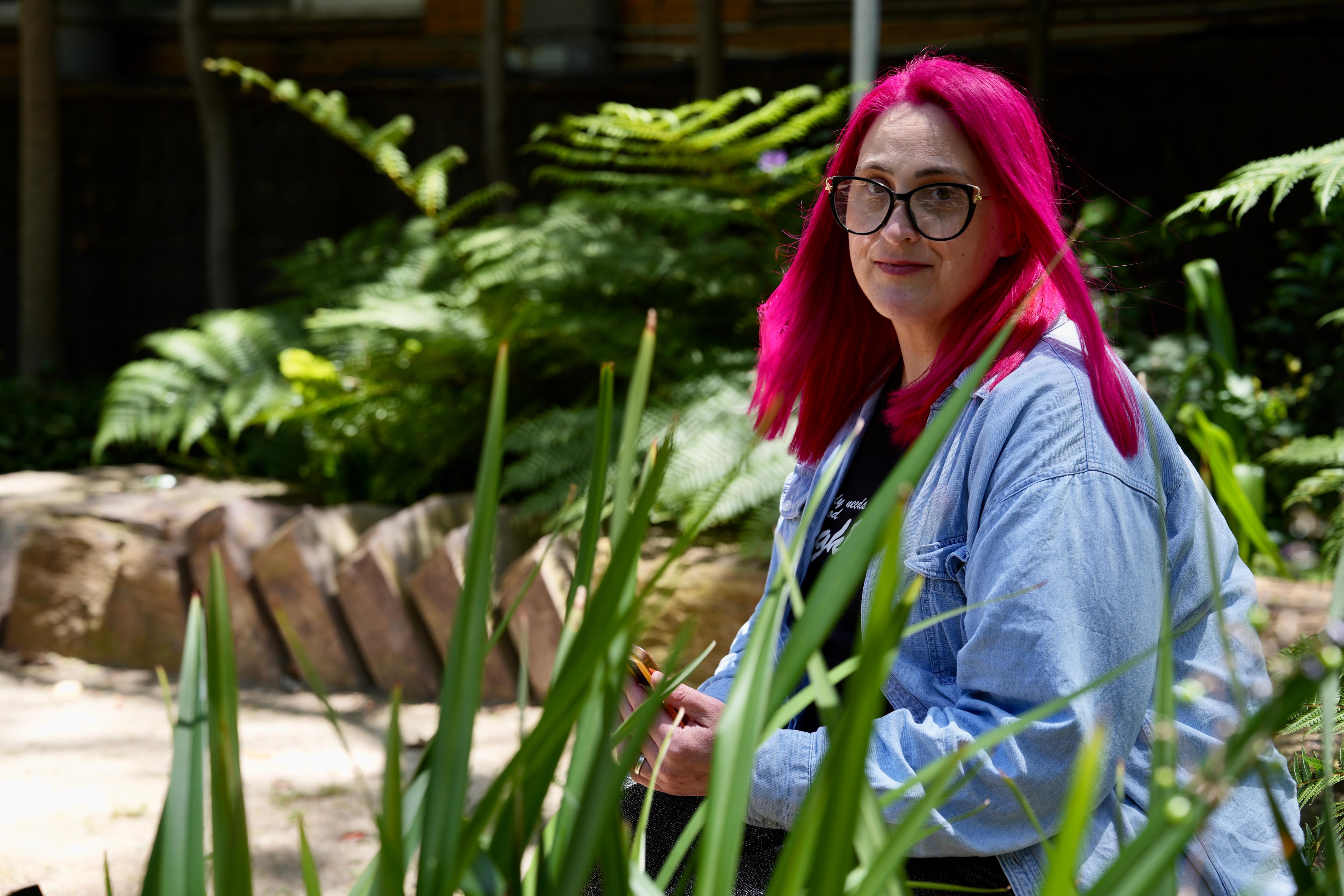 CJ tiene el pelo rosado y está sentado afuera detrás de algo de vegetación.