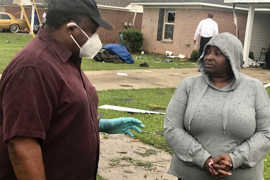 A man in a black cap and face mask and gloves talks to a woman in a grey hoodie with debris on her front lawn.