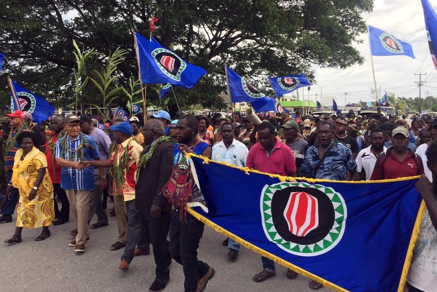 Bougainville President John Momis, in a blue shirt, walks with a crowd of people carrying the flag of Bougainville.