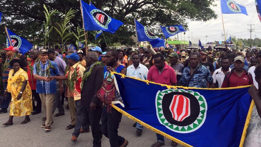 Bougainville President John Momis, in a blue shirt, walks with a crowd of people carrying the flag of Bougainville.