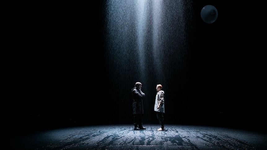 Colour production still of two characters standing face to face in the rain on stage in 2018 theatre production Saint Joan.