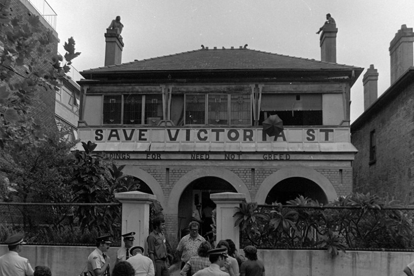 A black and white photo of people standing outside a house with Save Victoria Street painted on the facade.