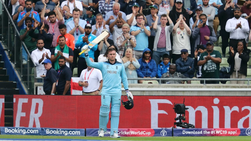 England batsman Jason Roy raises his bat and holds his helmet as cricket fans cheer behind him.