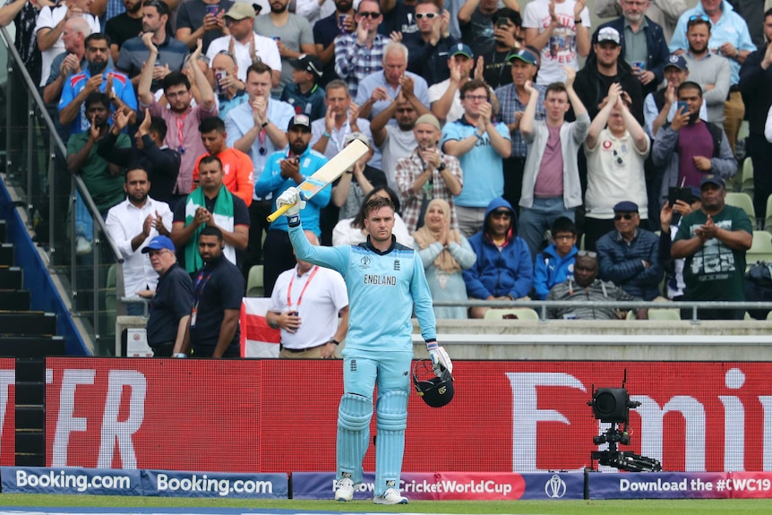 England batsman Jason Roy raises his bat and holds his helmet as cricket fans cheer behind him.