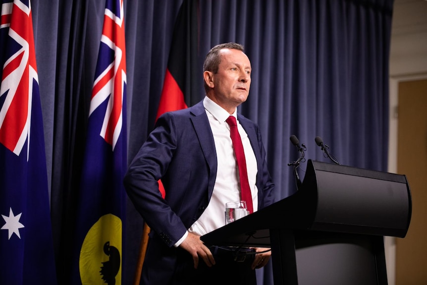WA Premier Mark McGowan at a lectern, hands on hips, with two flags behind him