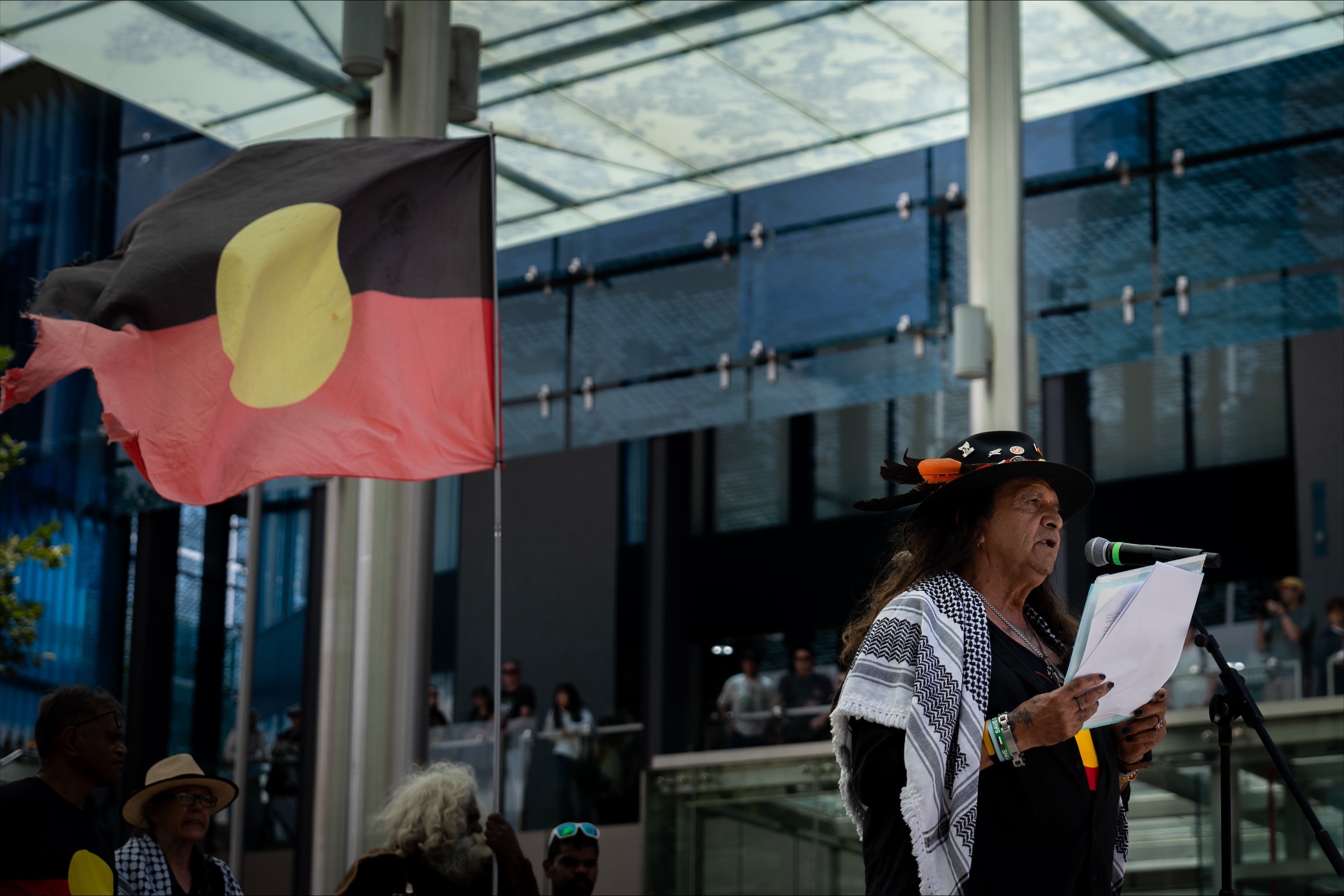 Noongar Elder Hedley Hayward speaks to the crowd with an Aboriginal flag behind.