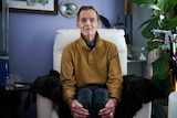 Paull Garrett sits on an armchair in his home, he holds his legs with his hands.