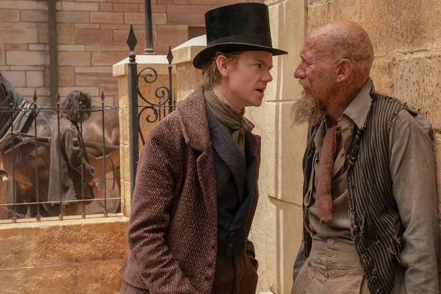 A young man in a top hat and period dress speaks tersely with an older man in a dishevelled state.