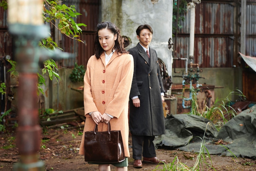 Still from a period drama showing a young woman and man standing outside in front of a building with some greenery.