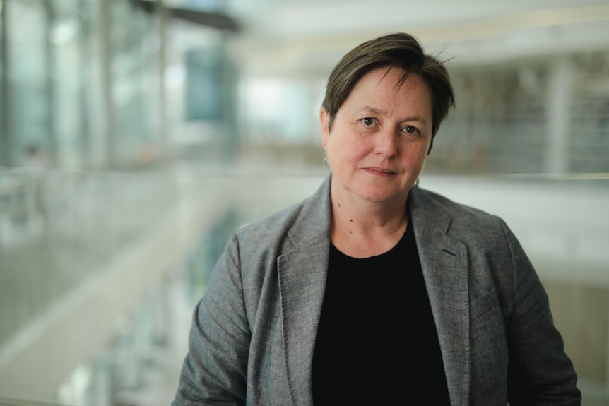 A woman stands inside a modern office building looking at the camera with a neutral expression.