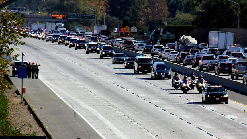 A line of dozens of police cars and motorbikes follow a black hearse on a highway.