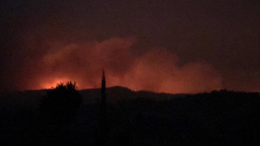 The orange glow of a fire above a hill at night.