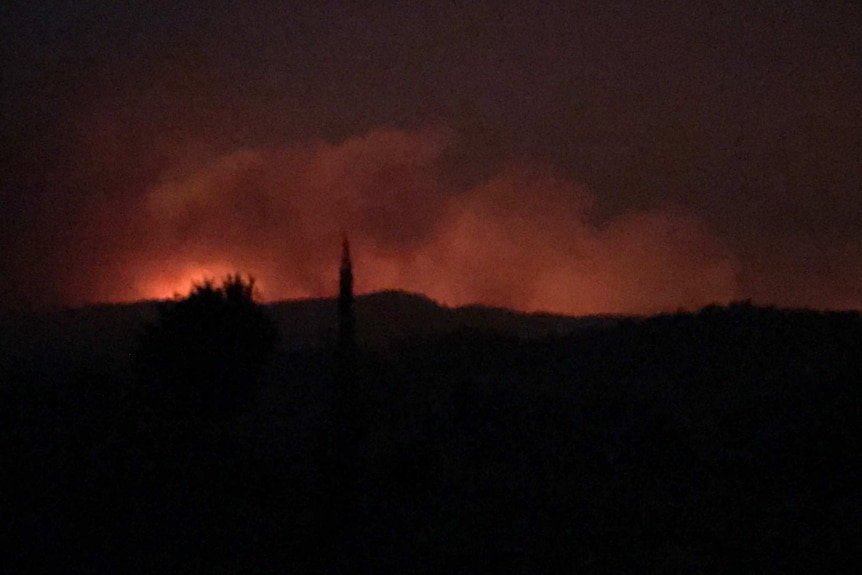 The orange glow of a fire above a hill at night.