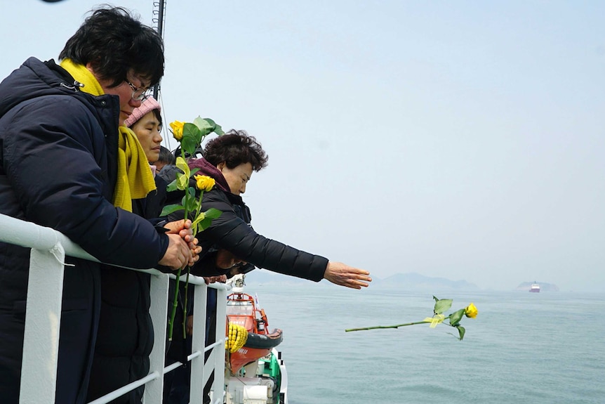 Grieving woman throws yellow rose into the ocean alongside other grieving relatives