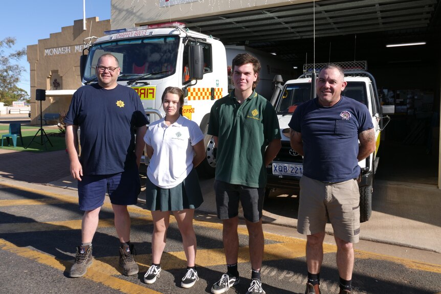 Four volunteers stand outside a fire station in front of a fire truck.