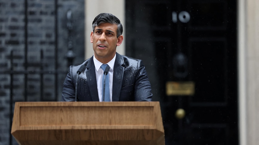 Rishi Sunak stands behind a wooden lectern in the street speaking in the rain