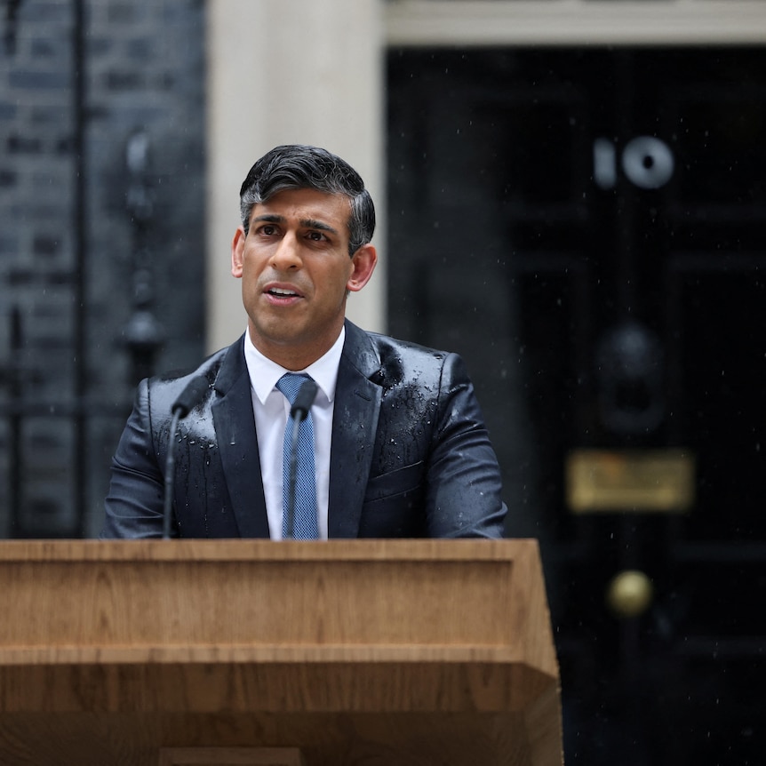 Rishi Sunak stands behind a wooden lectern in the street speaking in the rain