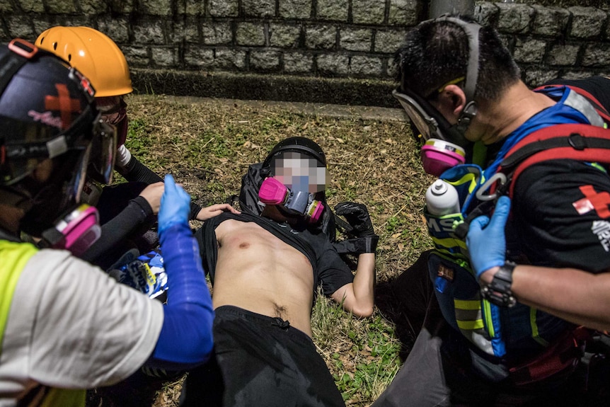 A protester lies on the ground wearing a facemask and surrounded by others offering medical assistance. His shirt is lifted up.