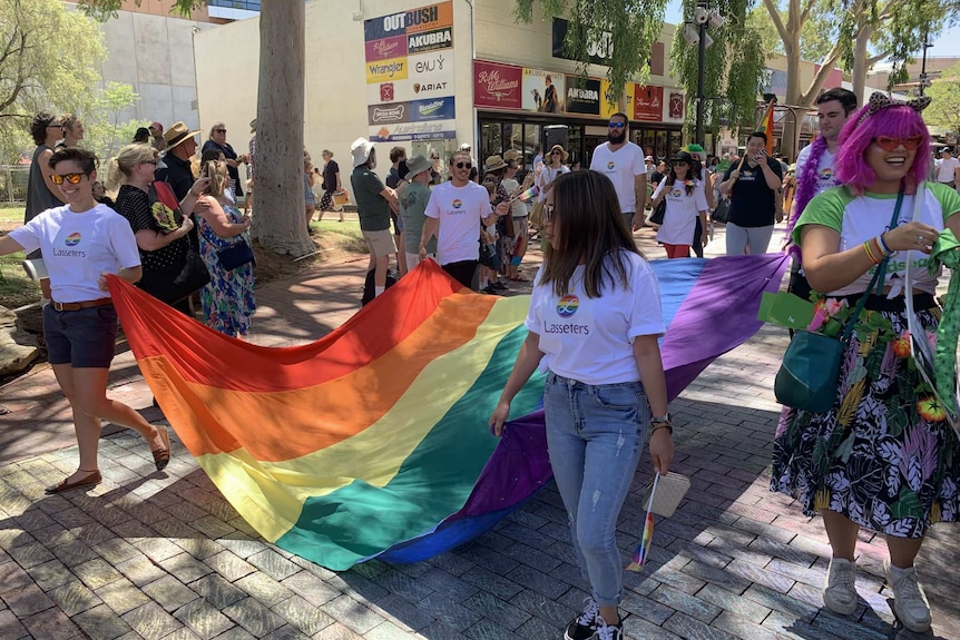 Two women and two men carry the corners of a rainbow flag down a mall while other people stand to the sides watching the parade.