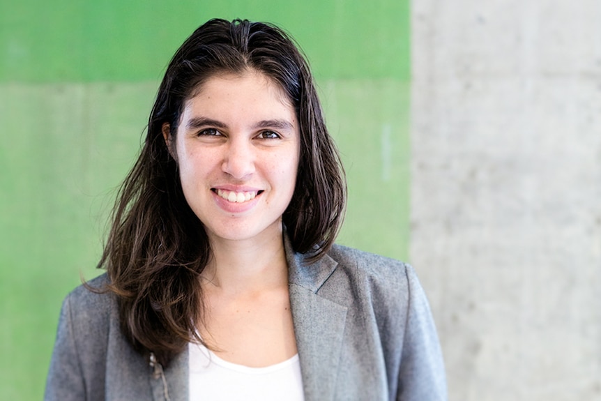 Colour photo of writer Ellen van Neerven smiling standing in front of green and grey concrete wall.