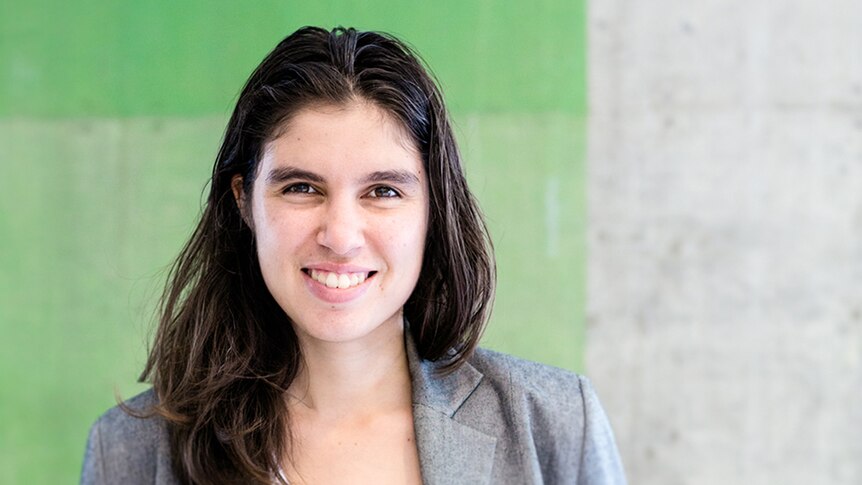 Colour photo of writer Ellen van Neerven smiling standing in front of green and grey concrete wall.