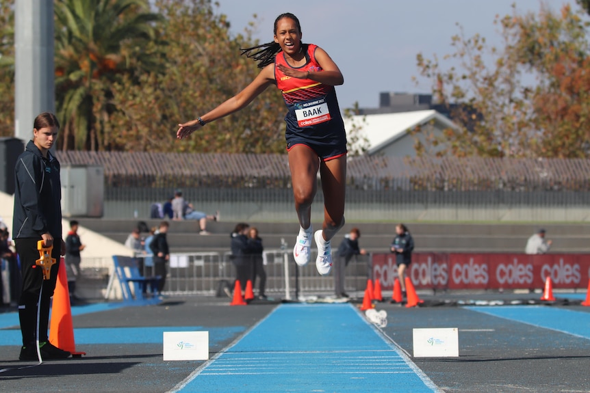 A girl with long brown hair bounds down an athletics track and jumps toward a long-jump sandpit