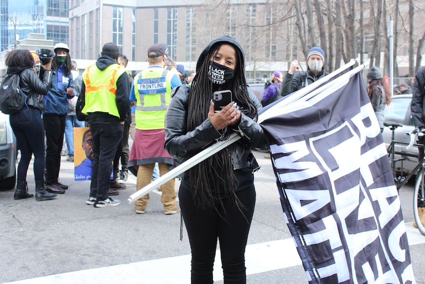 A woman wearing a black shirt and pants and a mask holds a sign wth Black Lives Matter written on it.